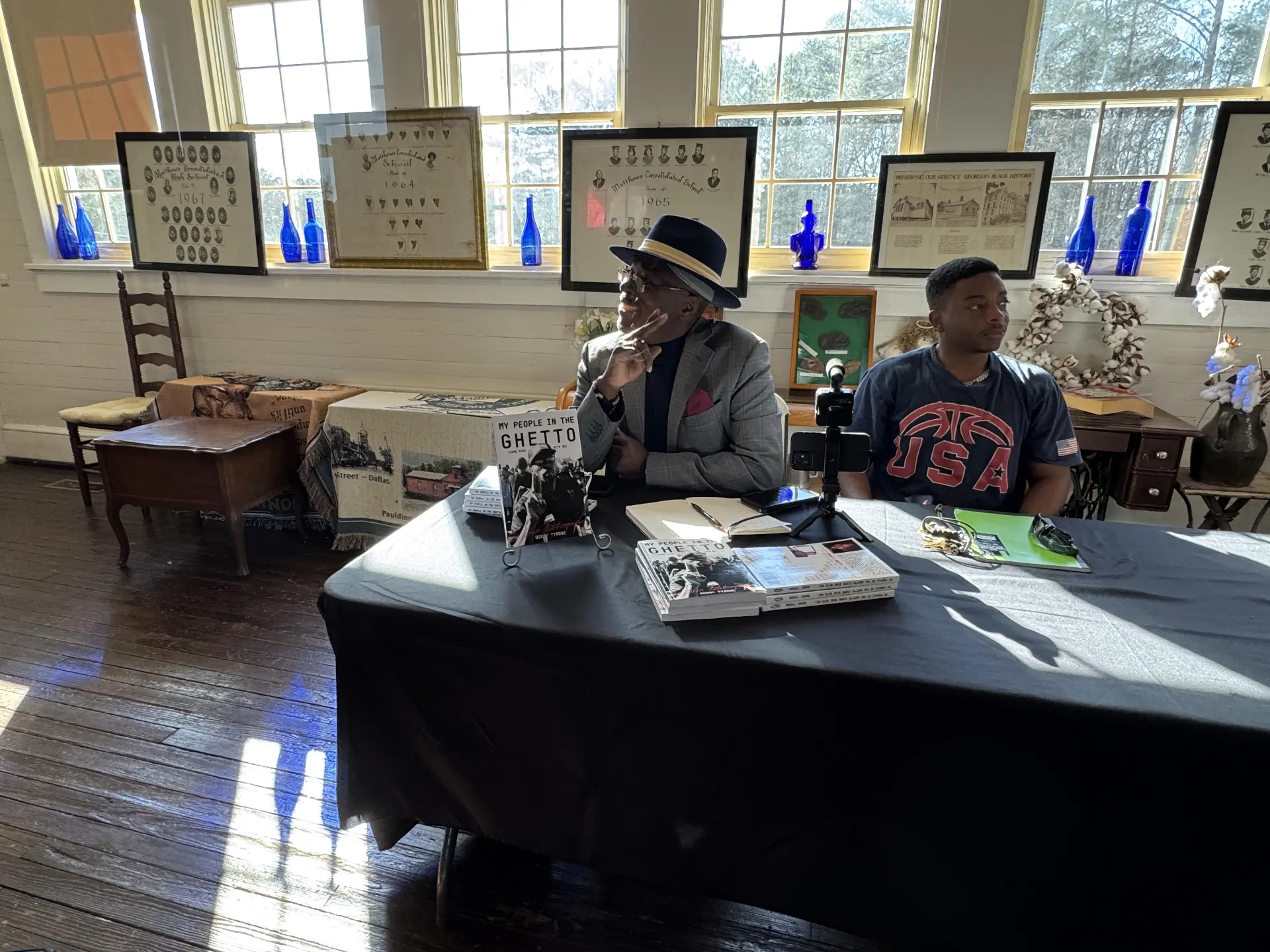An author in a hat sits at a signing table stacked with copies of My People in the Ghetto alongside a young guest.