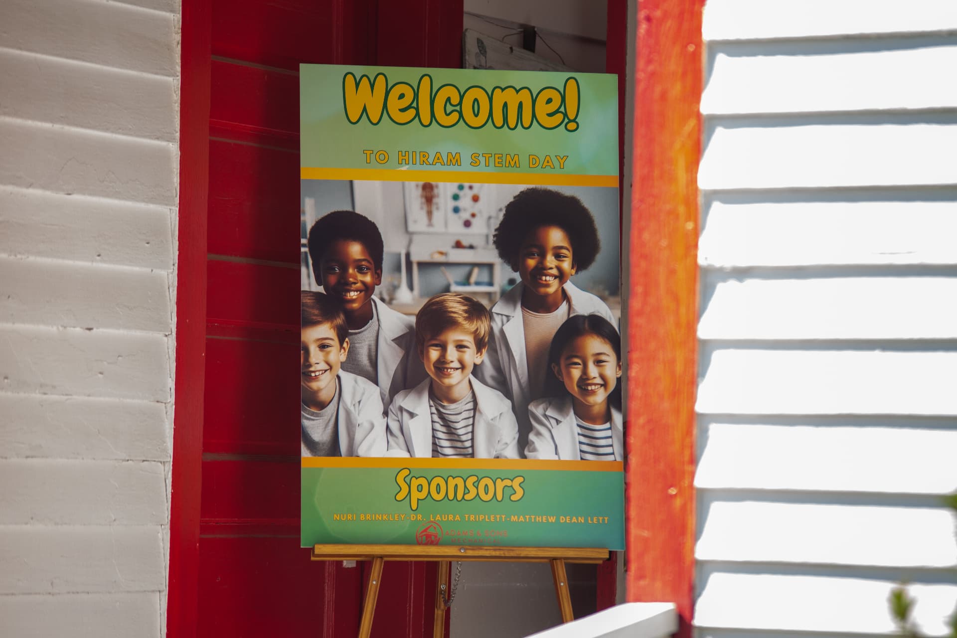 A Welcome to Hiram STEM Day sign with sponsor names stands on an easel by the red door of the schoolhouse.