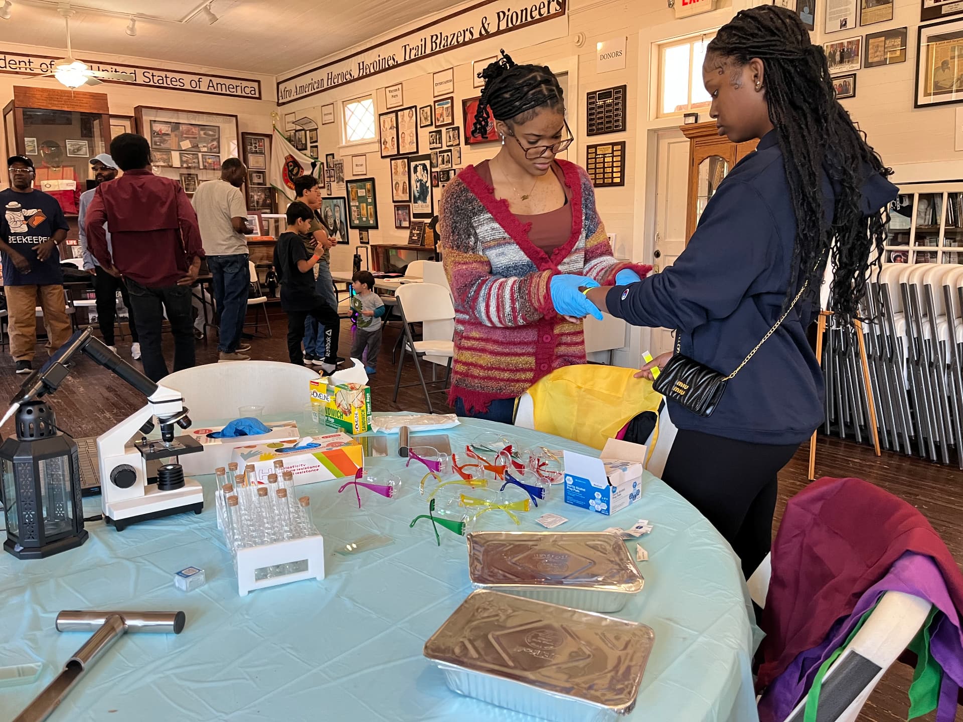 A volunteer helps a student pull on blue gloves beside a table of safety goggles and a microscope during STEM Day.