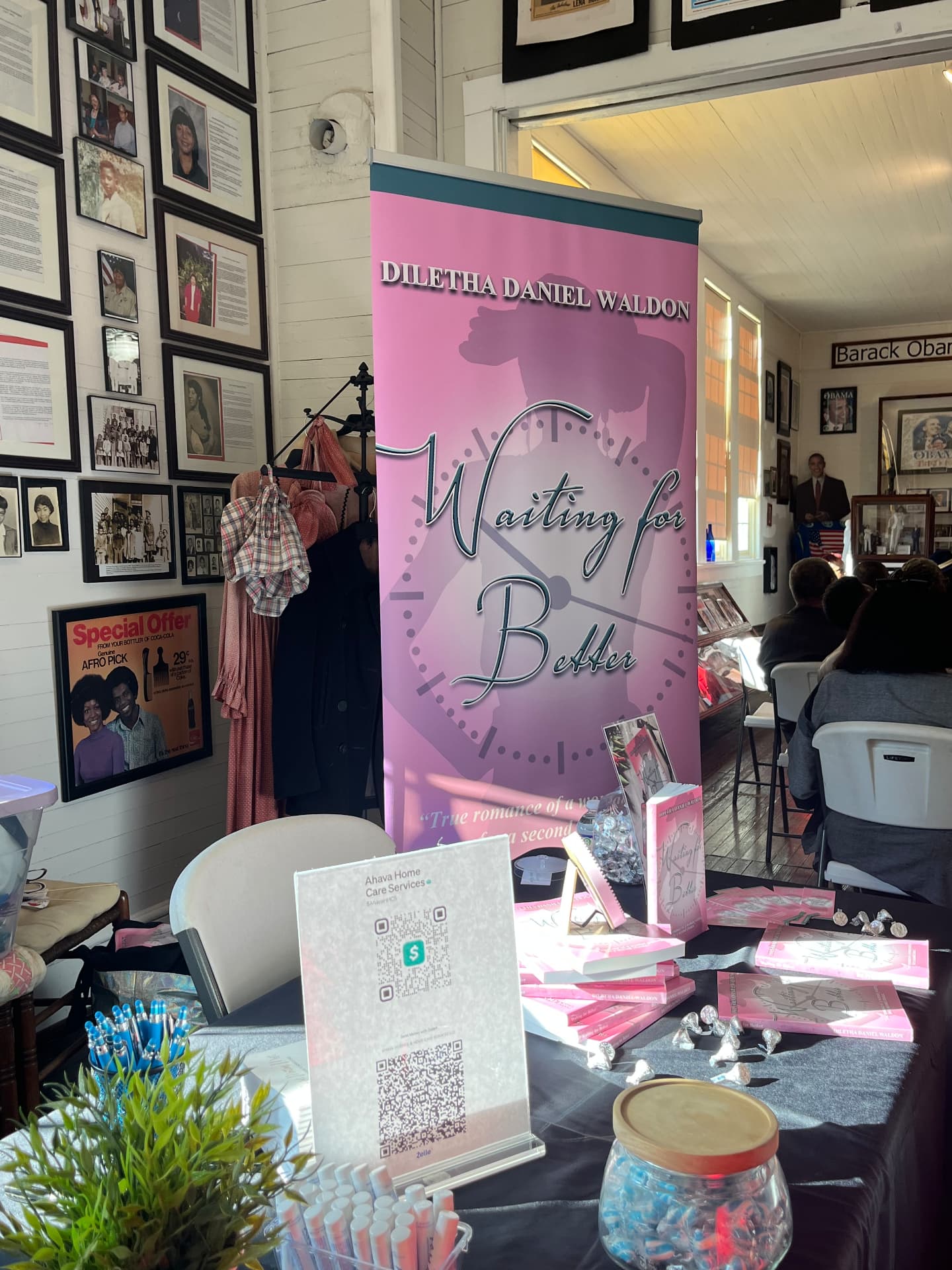 A pink Diletha Daniel Waldon Waiting for Better banner stands beside a vendor table with books and treats.