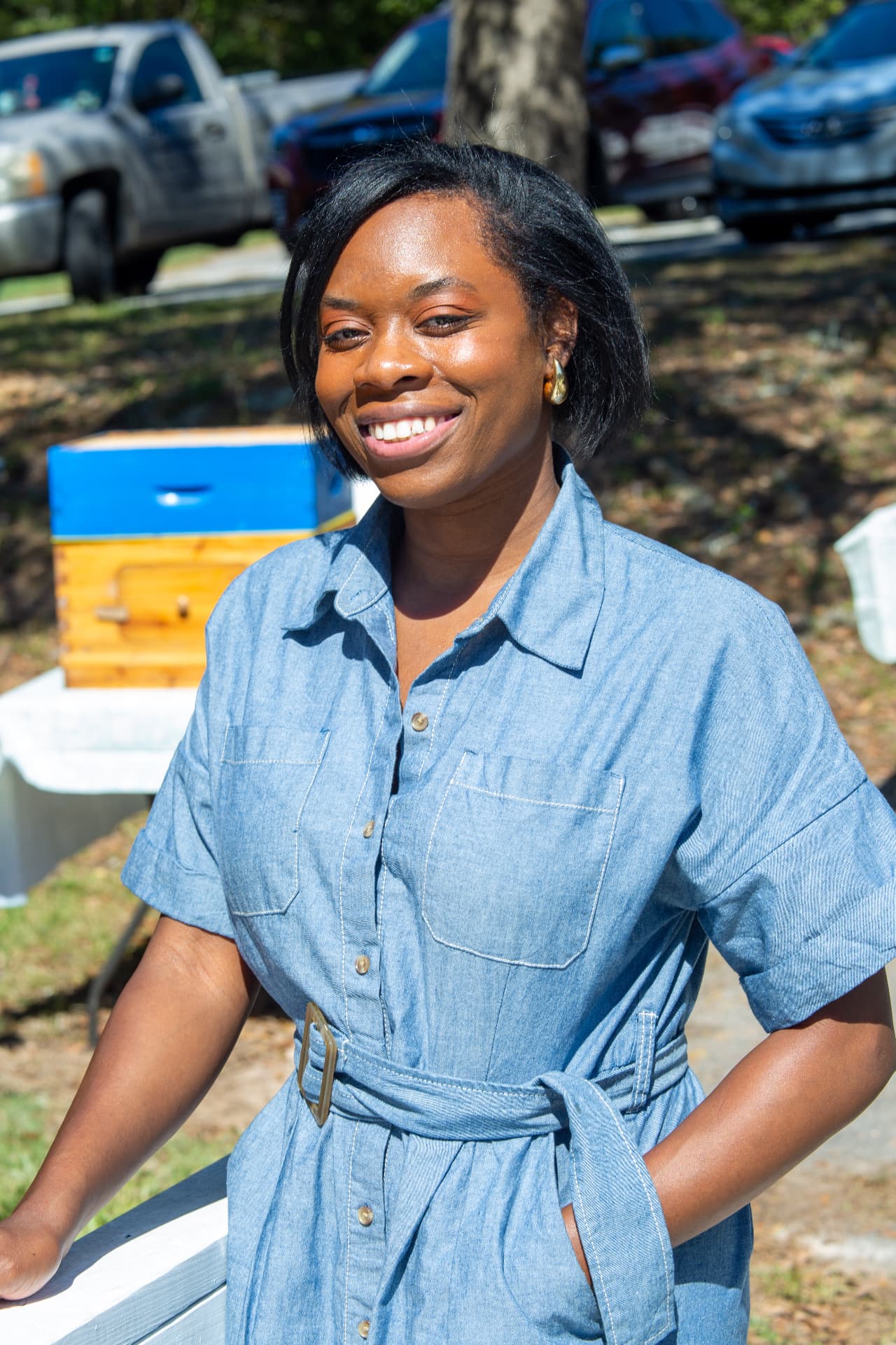 A smiling organizer in a denim dress poses near the museum porch and beehives.