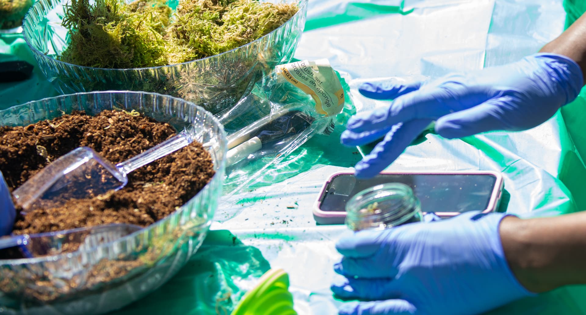 Gloved hands scoop soil and moss from bowls on an activity table during the STEM Day terrarium session.