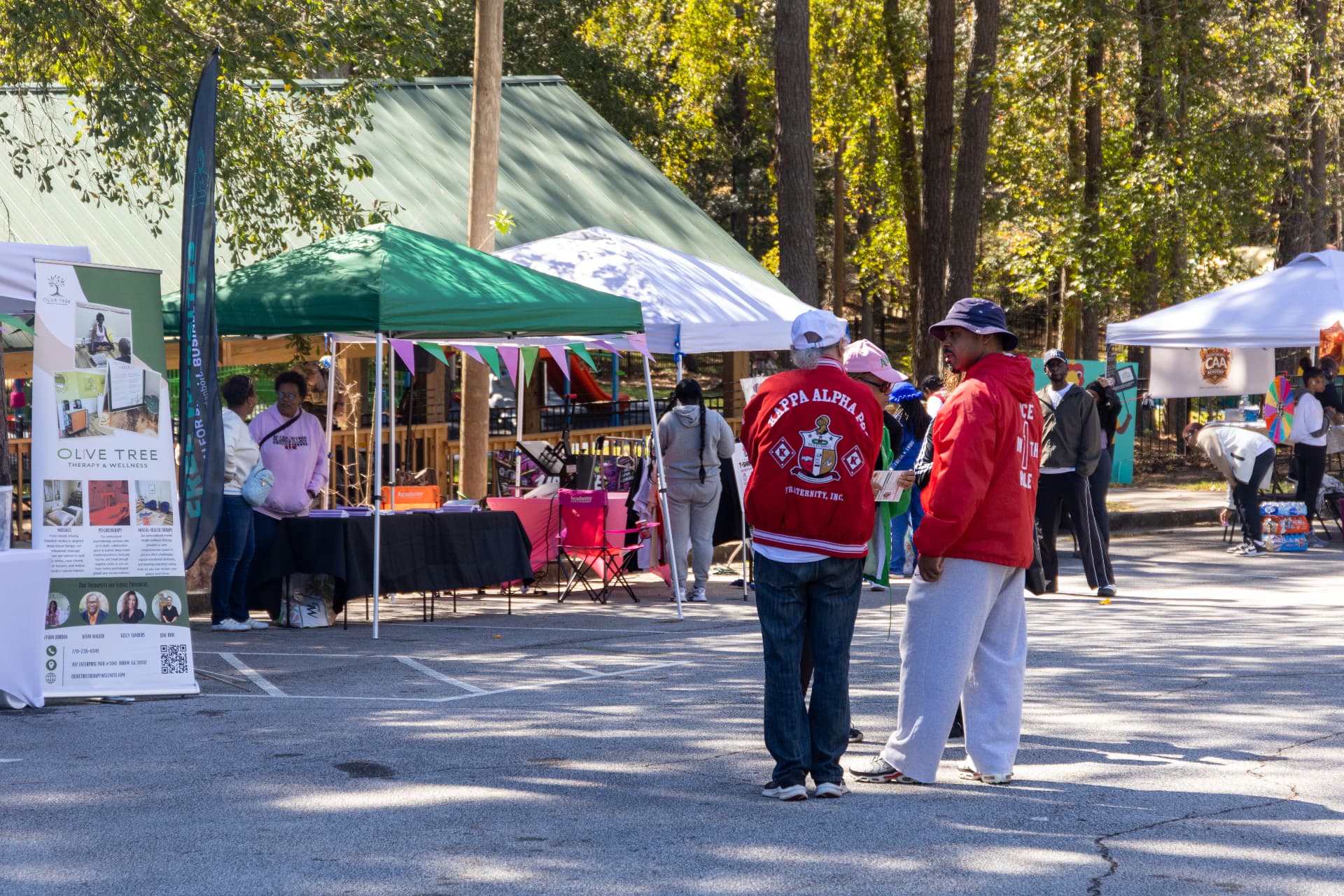 Guests browse vendor tents including Olive Tree Therapy & Wellness while fraternity members chat in the parking lot.