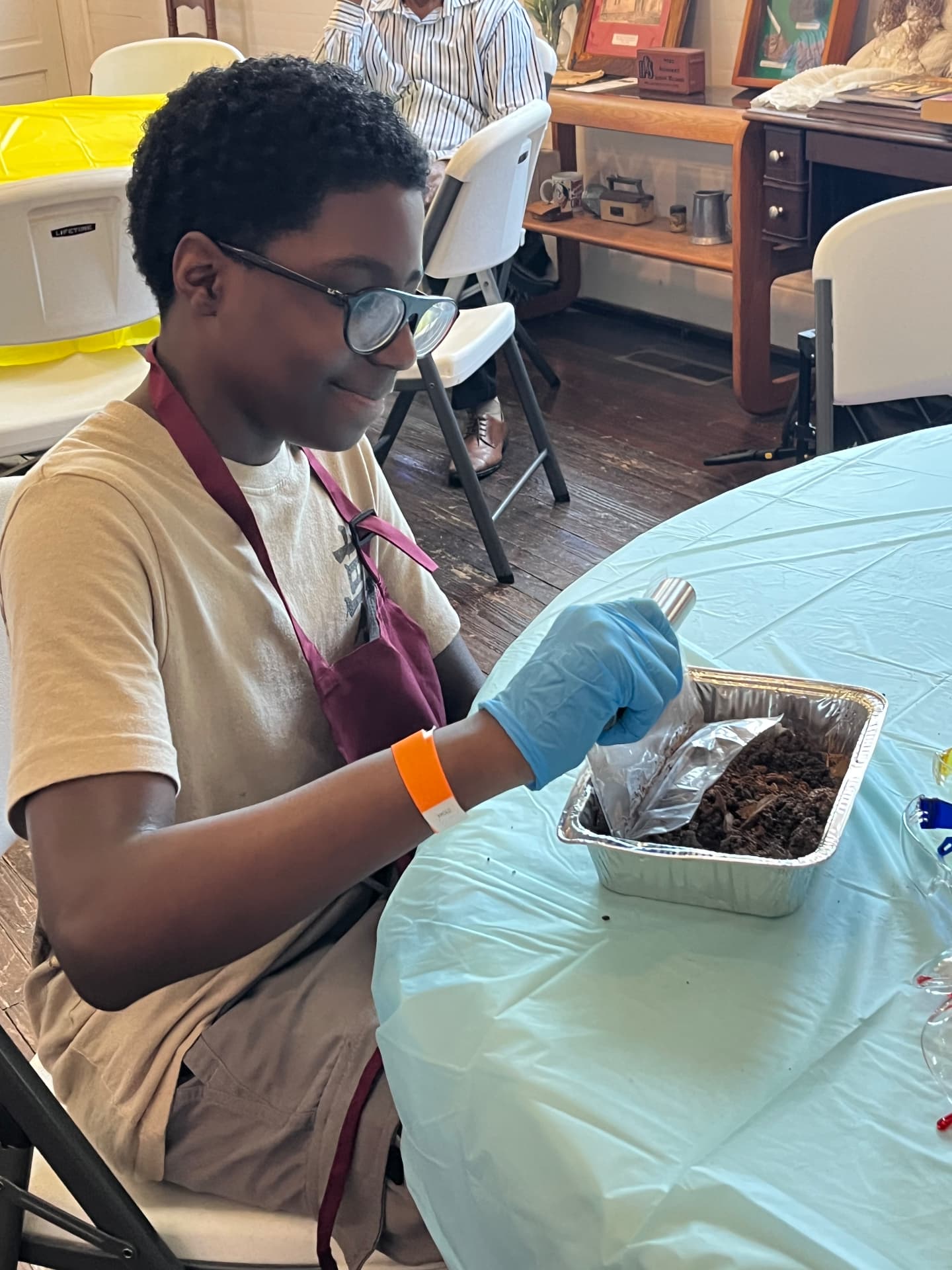 A smiling student in an apron and gloves scoops soil into an aluminum pan for a STEM Day activity.