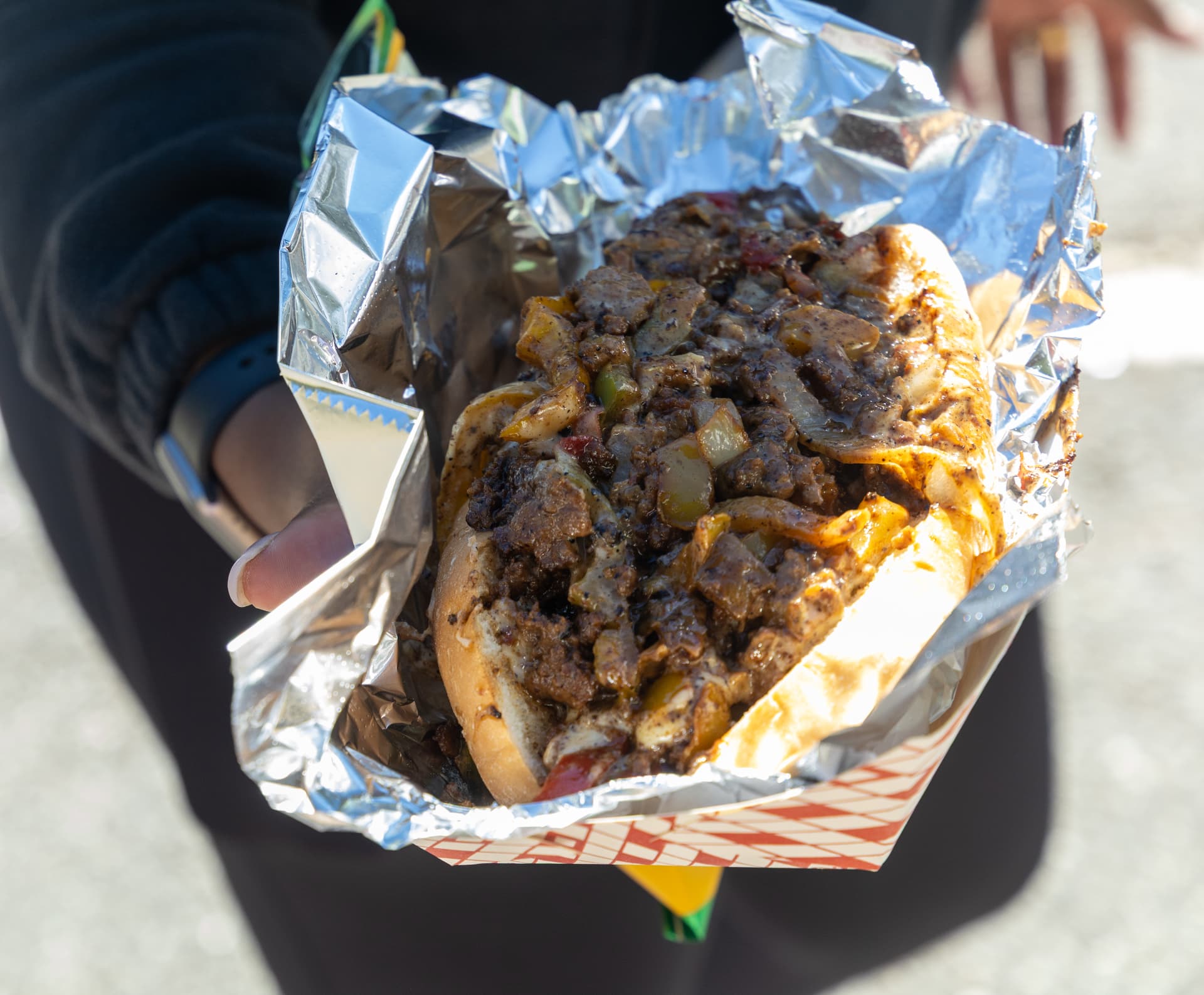 A loaded cheesesteak sandwich with peppers and onions served in foil from a block party food vendor.