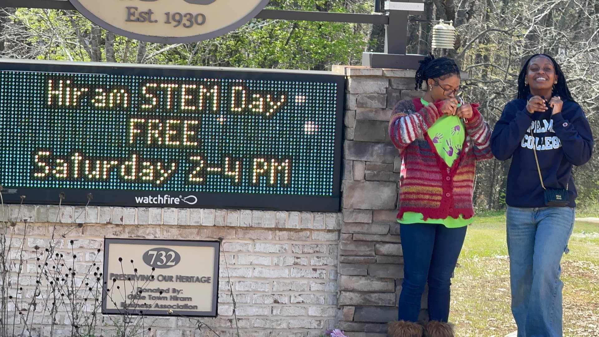 Two smiling students pose beside the museum's digital marquee announcing Hiram STEM Day, free Saturday 2-4 PM.