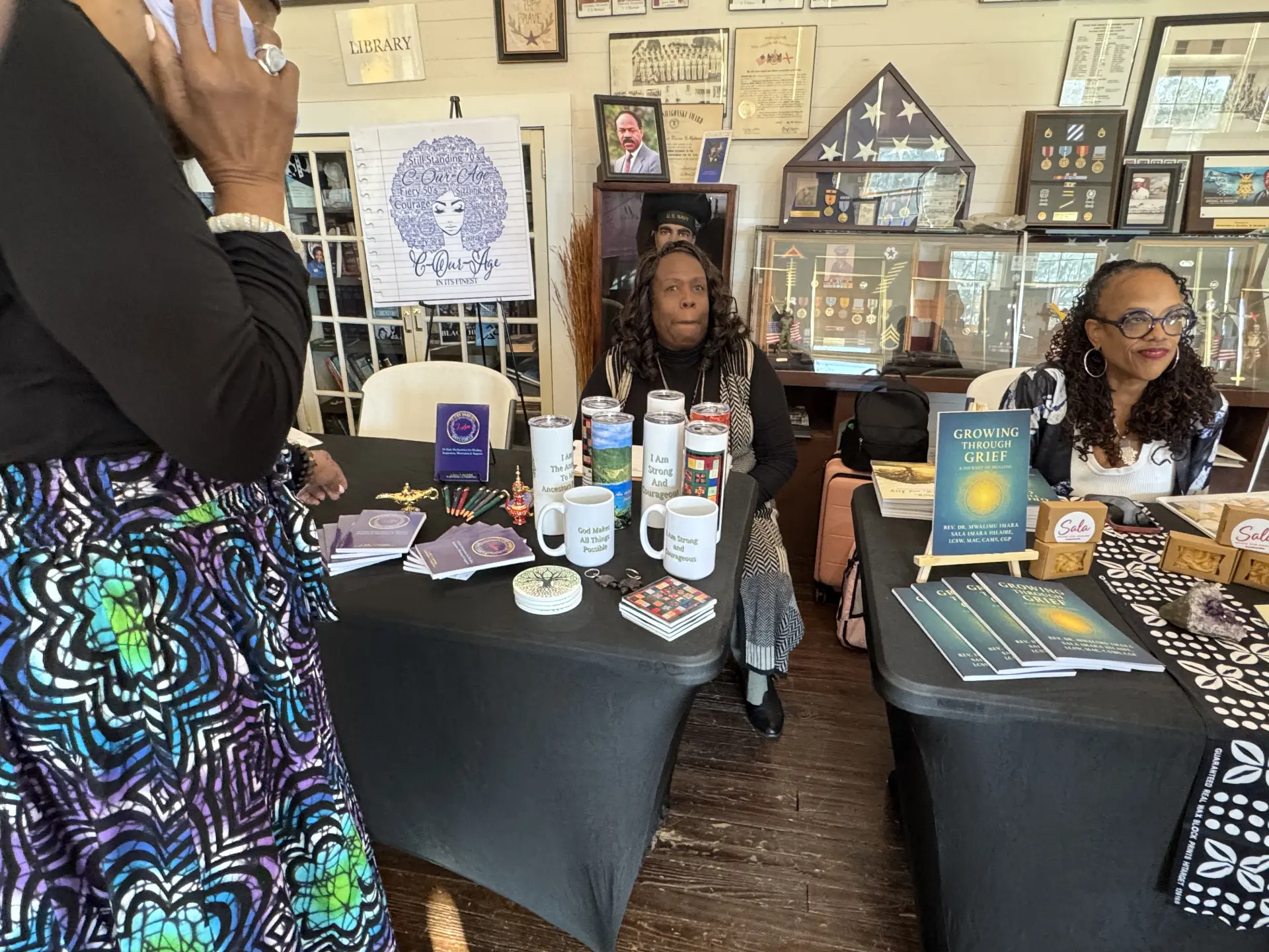 Vendor tables display purple-covered devotionals, mugs and tumblers beneath an C-Our-Age banner in the museum.