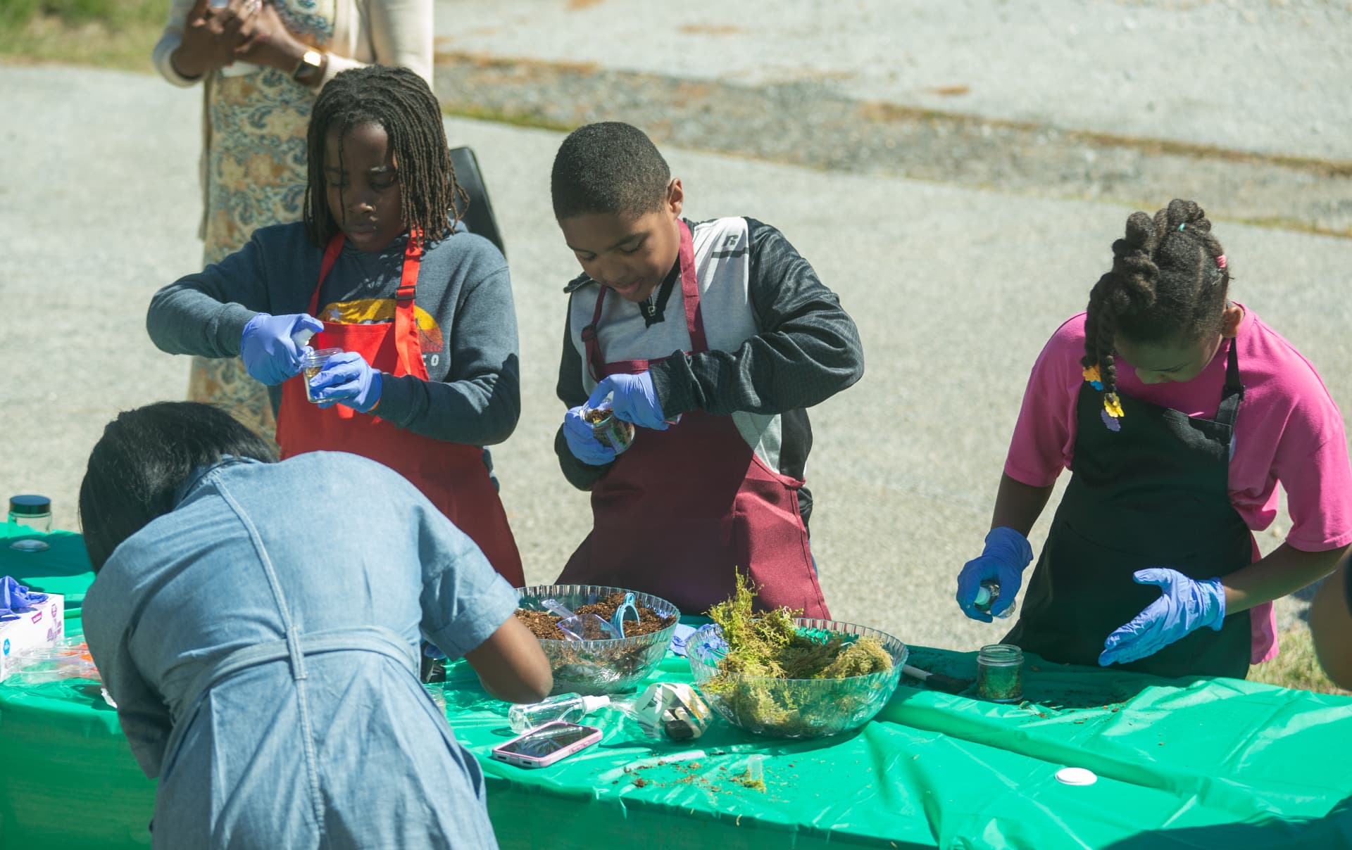 Children in aprons fill jars with soil and moss at the outdoor STEM Day terrarium table.