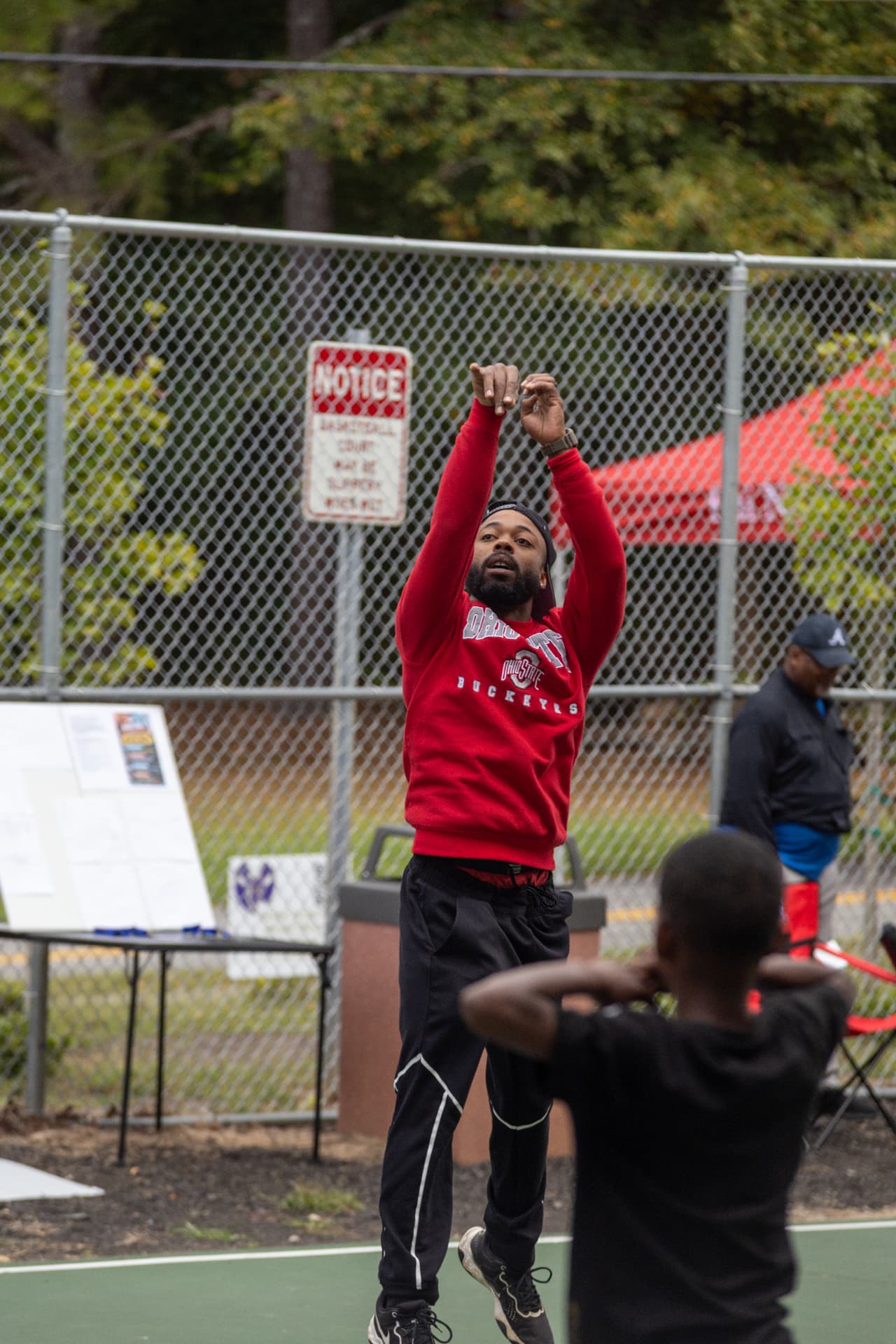 A man in a red Ohio State Buckeyes sweatshirt shoots a basketball on the outdoor court during the block party.