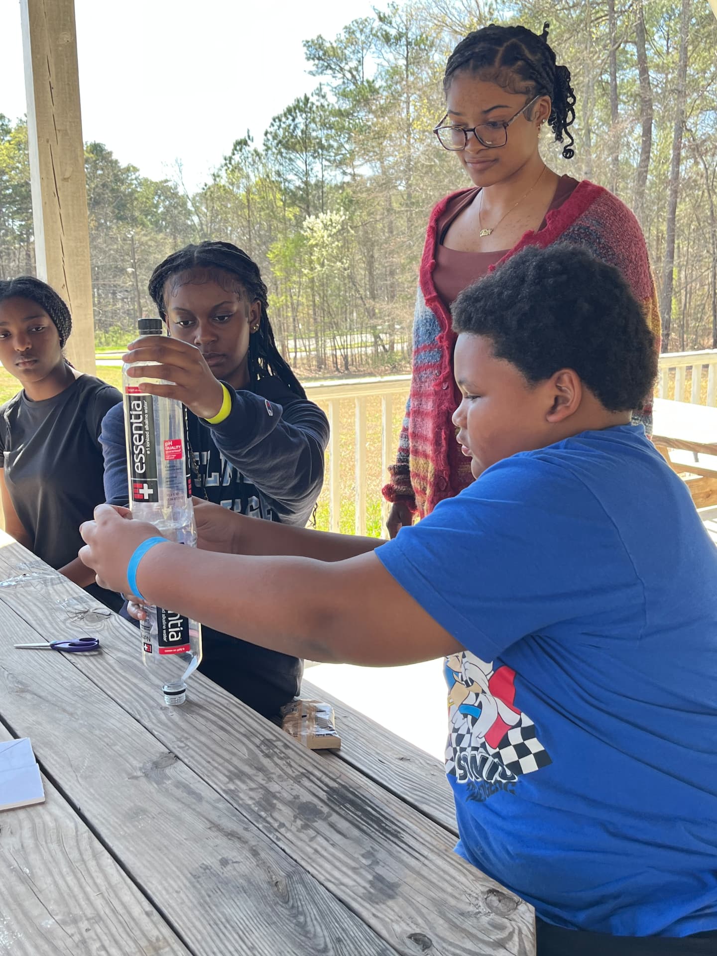 Students cut a plastic water bottle with scissors as they build their STEM Day rocket experiment under the pavilion.