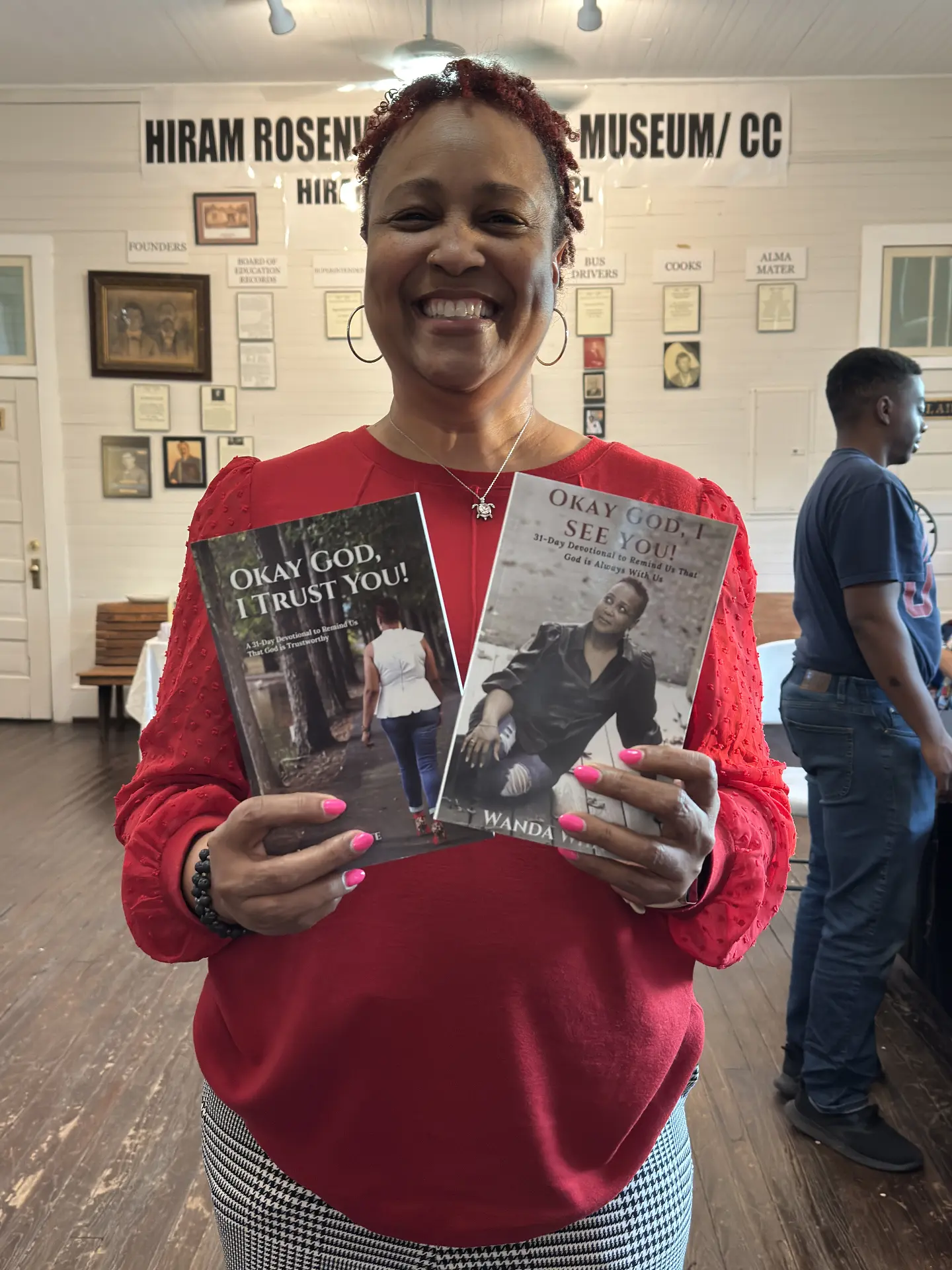 A smiling author in red holds her devotional books Okay God, I Trust You! and Okay God, I See You! inside the museum.