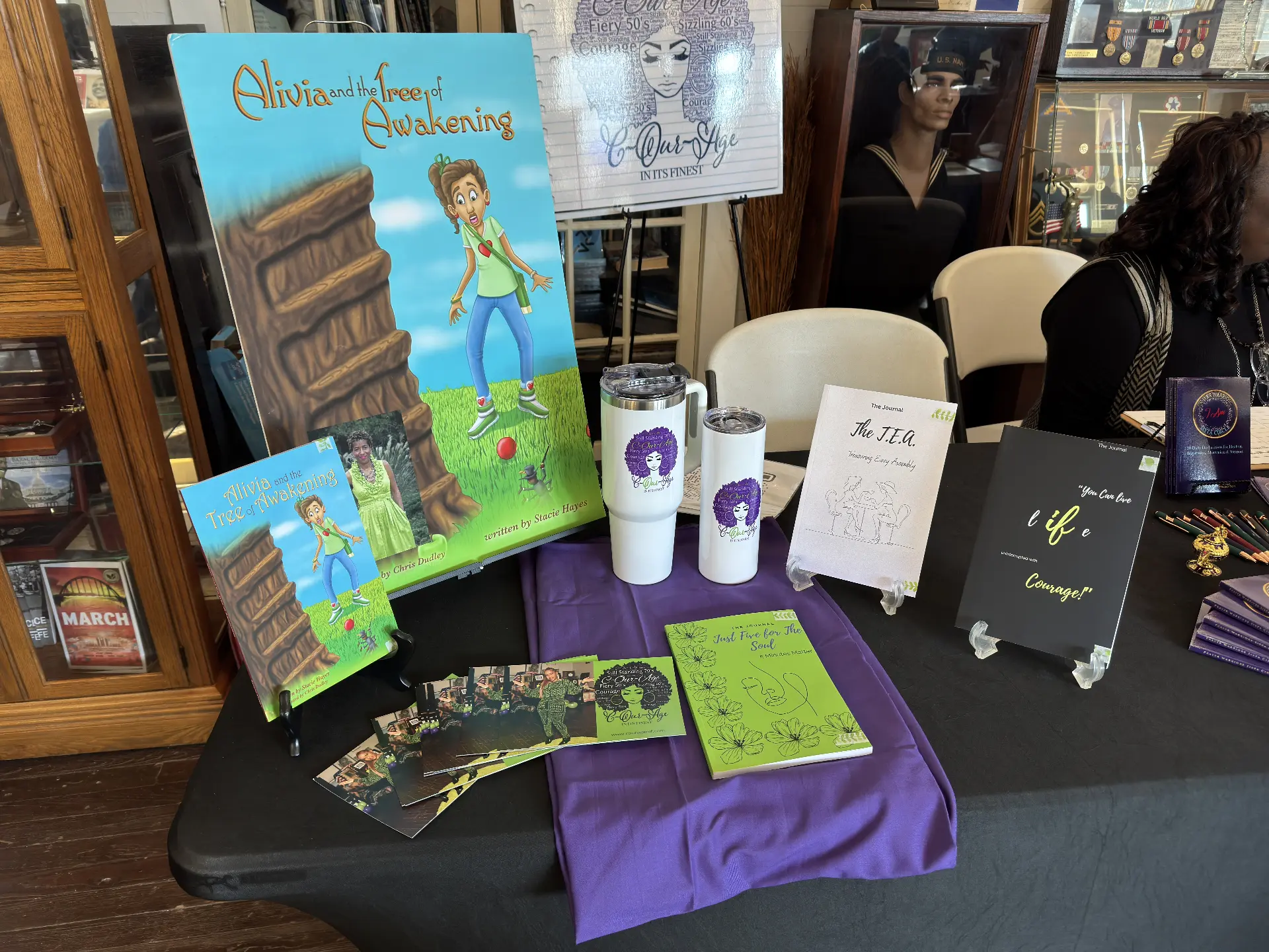 A vendor table featuring the children's book Alivia and the Tree of Awakening alongside branded tumblers and journals.