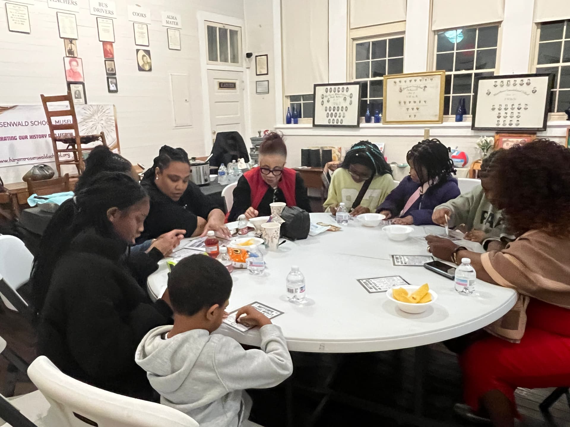 Guests of all ages gather around a table playing bingo beneath the museum's classroom displays and framed class portraits.