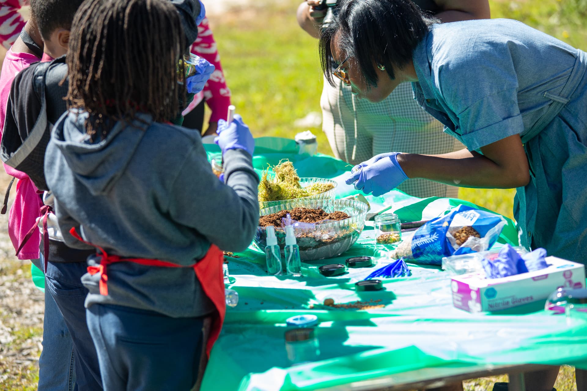 An instructor helps children fill glass jars with soil and moss at the STEM Day terrarium activity.
