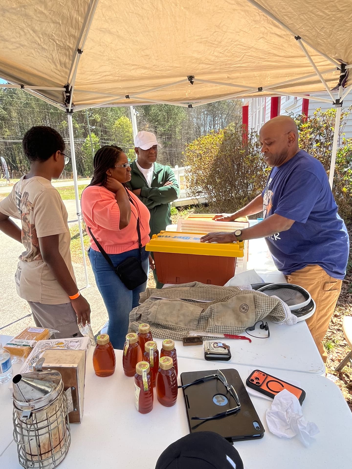 A beekeeper shows a bee box and honey jars to visitors around a table of supplies under the STEM Day tent outside the museum.