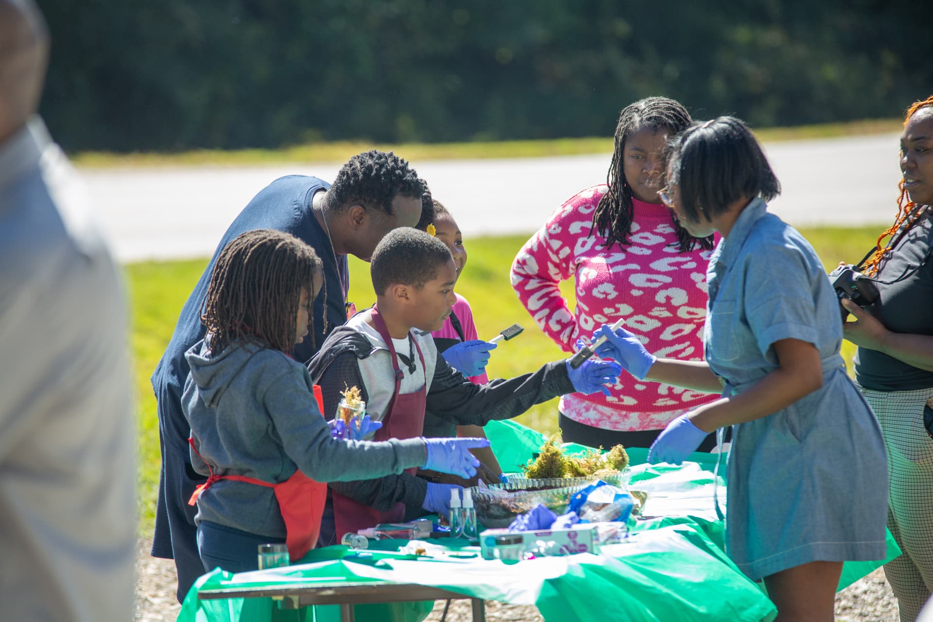 An instructor in gloves helps students in aprons build terrariums at an outdoor STEM Day table.