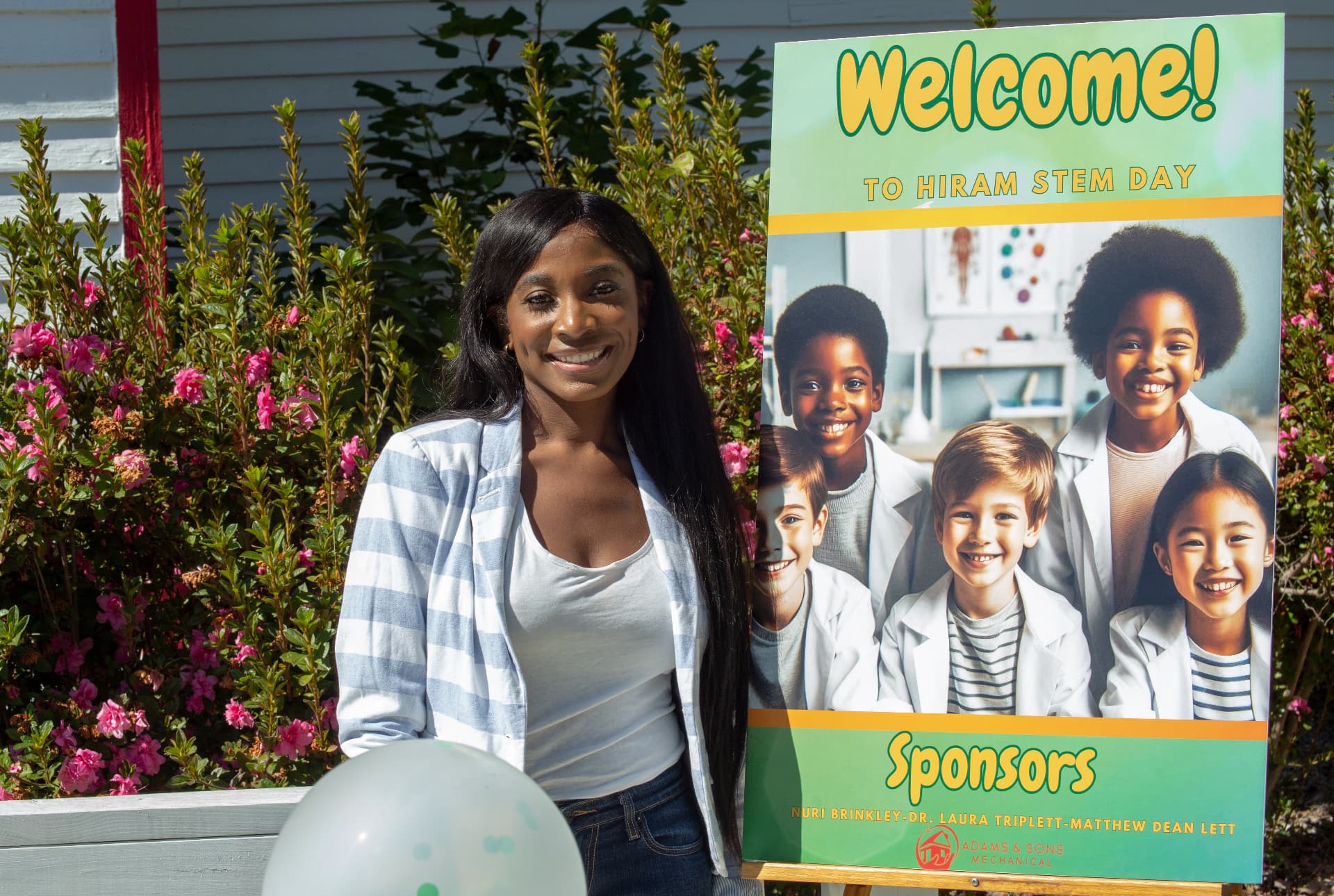 A STEM Day host poses next to the Welcome sign in front of blooming azaleas.