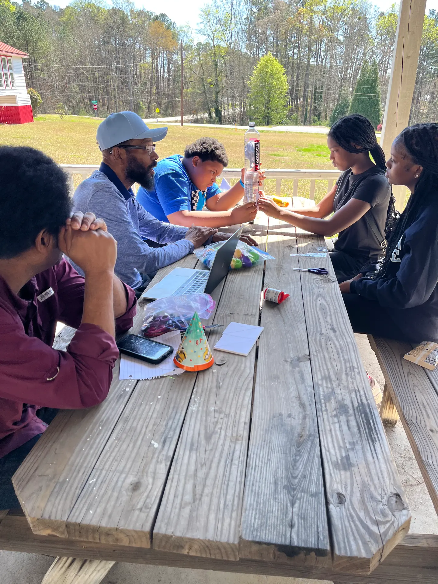 An instructor and students test a water-bottle experiment at a picnic table during STEM Day, with the schoolhouse in the background.