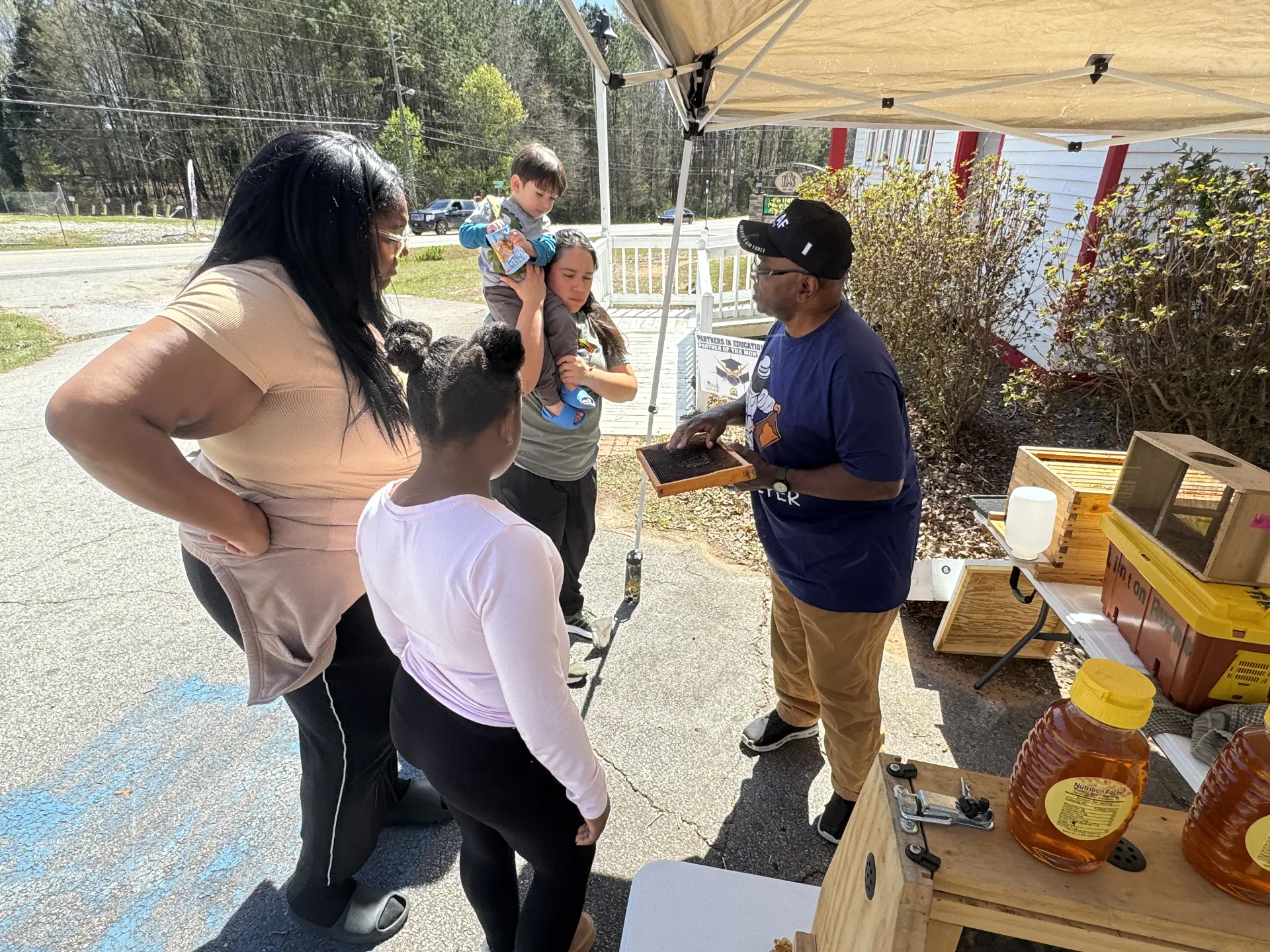 A beekeeper holds up a honeycomb frame to explain bees to a family and young child during STEM Day.