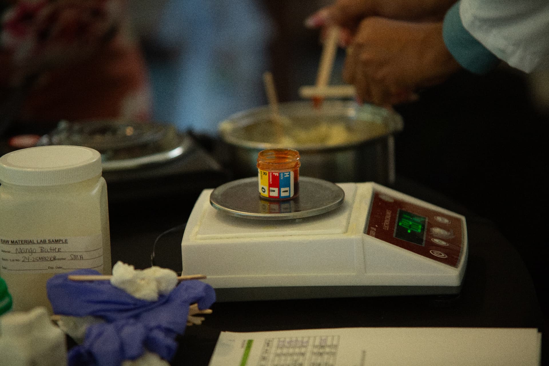A small jar of colored pigment sits on a digital scale while a scientist stirs a warm mixture during the STEM Day cosmetics lab.