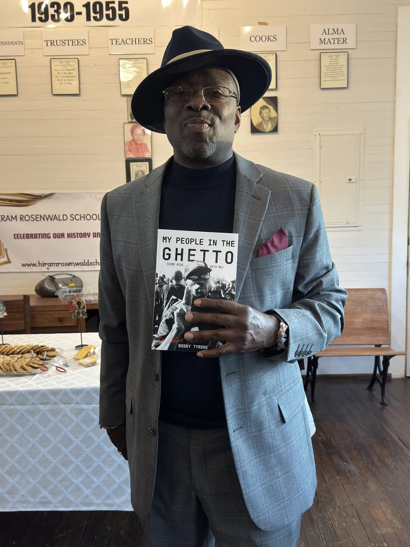 An author in a plaid suit holds his book My People in the Ghetto in front of the Hiram Rosenwald School celebration banner.