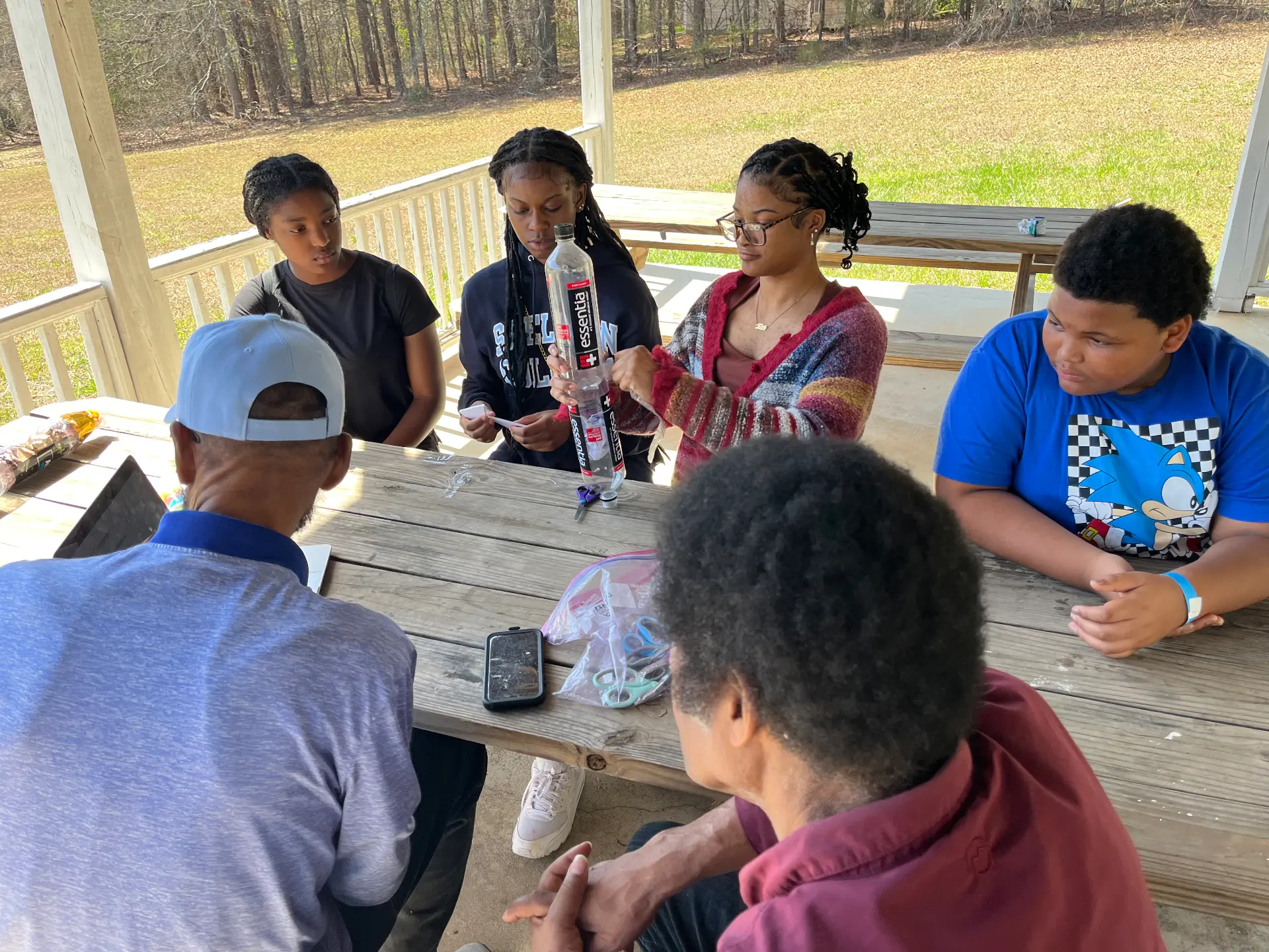 Students at a pavilion table hold up a plastic bottle as they prepare their STEM Day experiment.