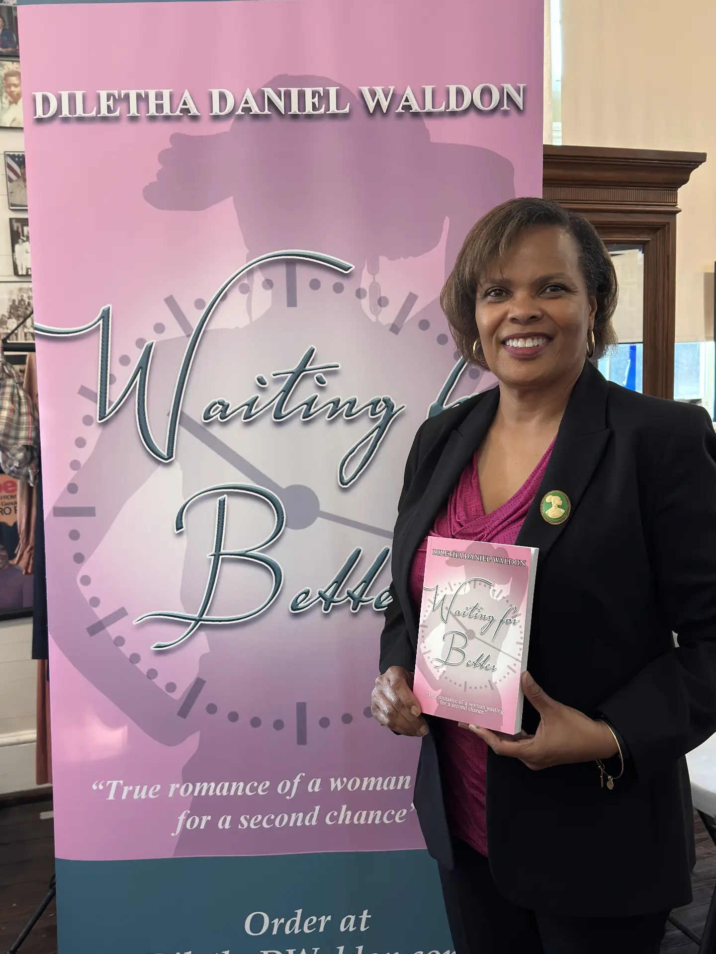 Author Diletha Daniel Waldon holds her novel Waiting for Better in front of its pink promotional banner.