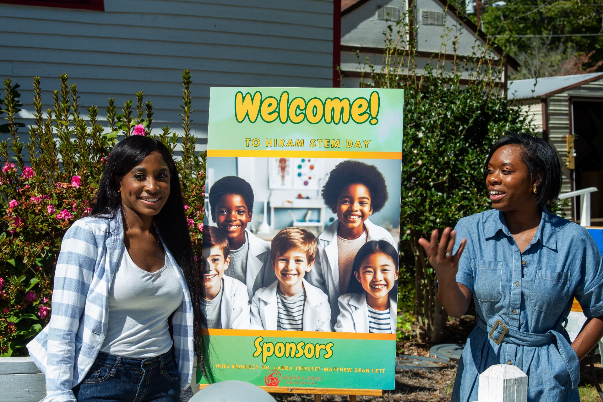 Two organizers smile beside the Welcome to Hiram STEM Day sponsor sign outside the schoolhouse.