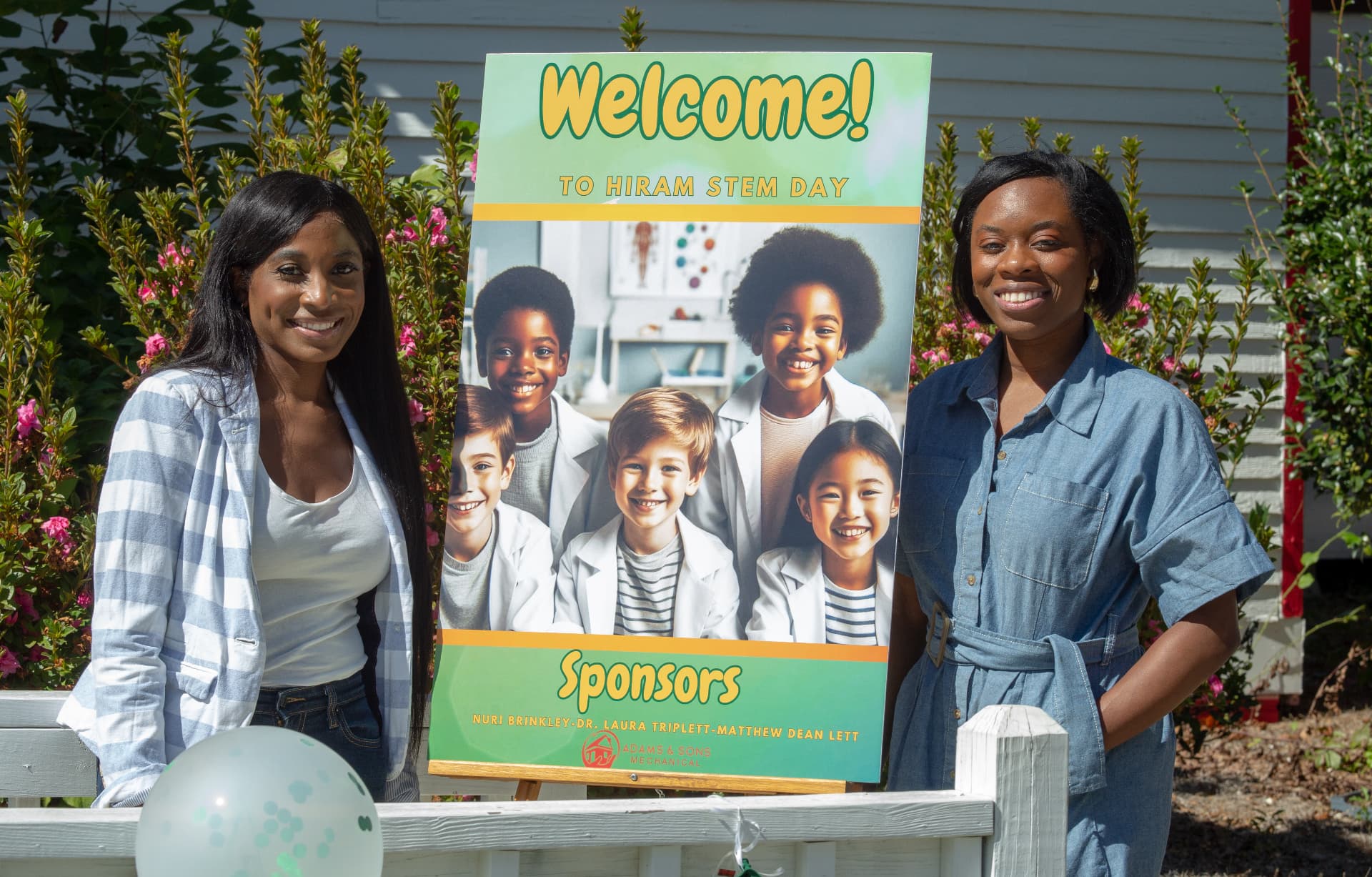 Hosts smile beside the STEM Day welcome sign listing event sponsors.