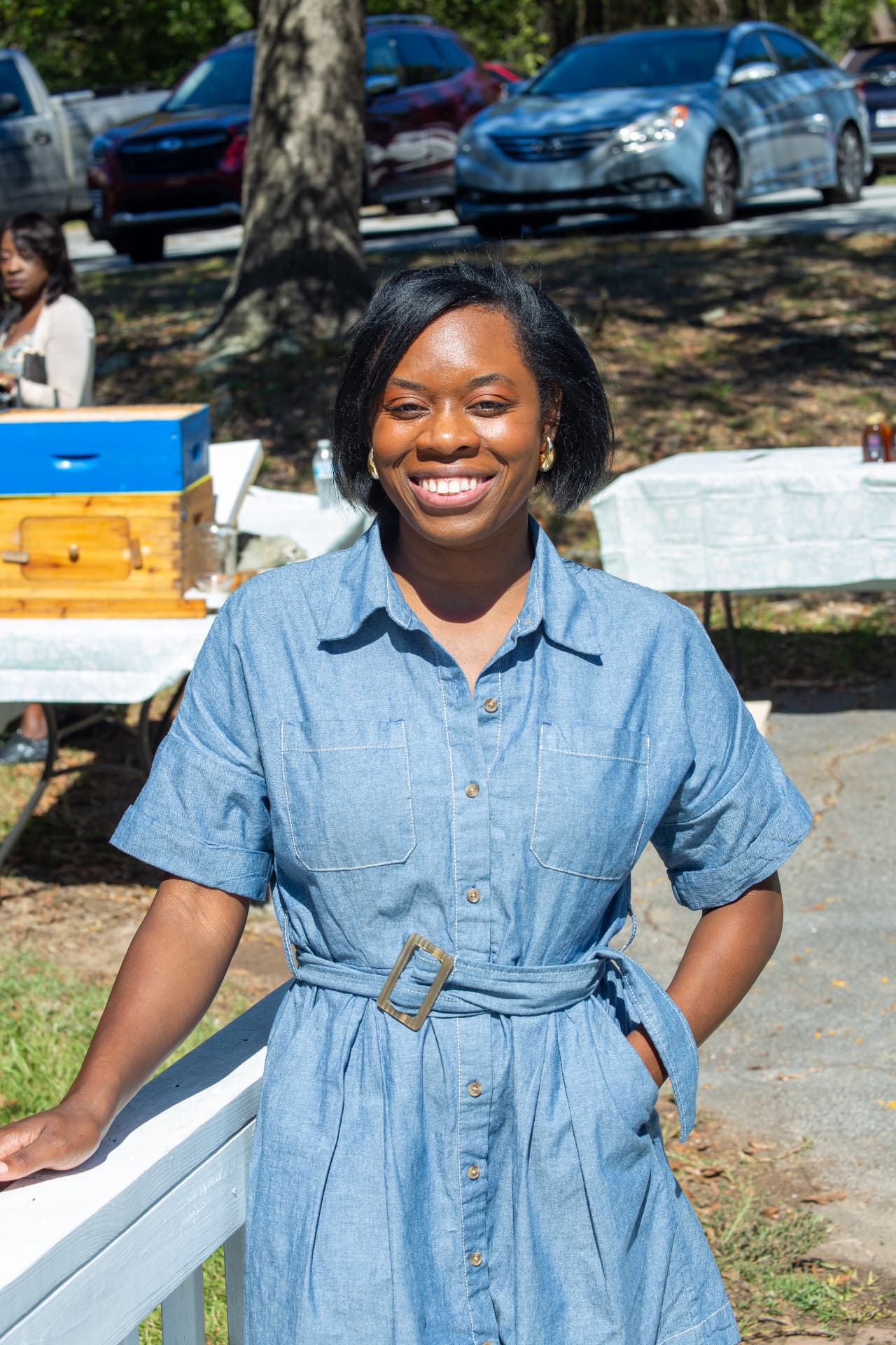 A smiling organizer in a denim shirt dress stands outdoors by the museum porch during STEM Day.
