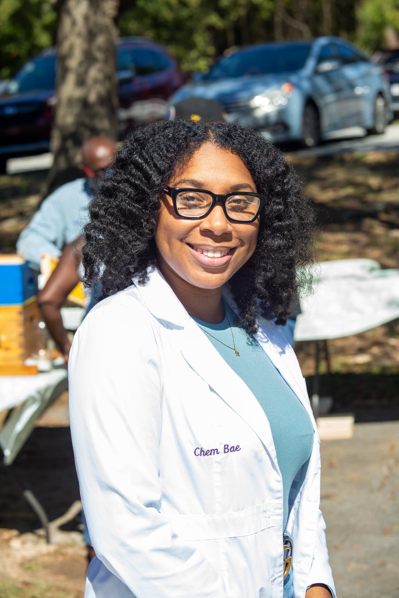 The guest scientist in her lab coat smiles outdoors near the beehives during STEM Day.