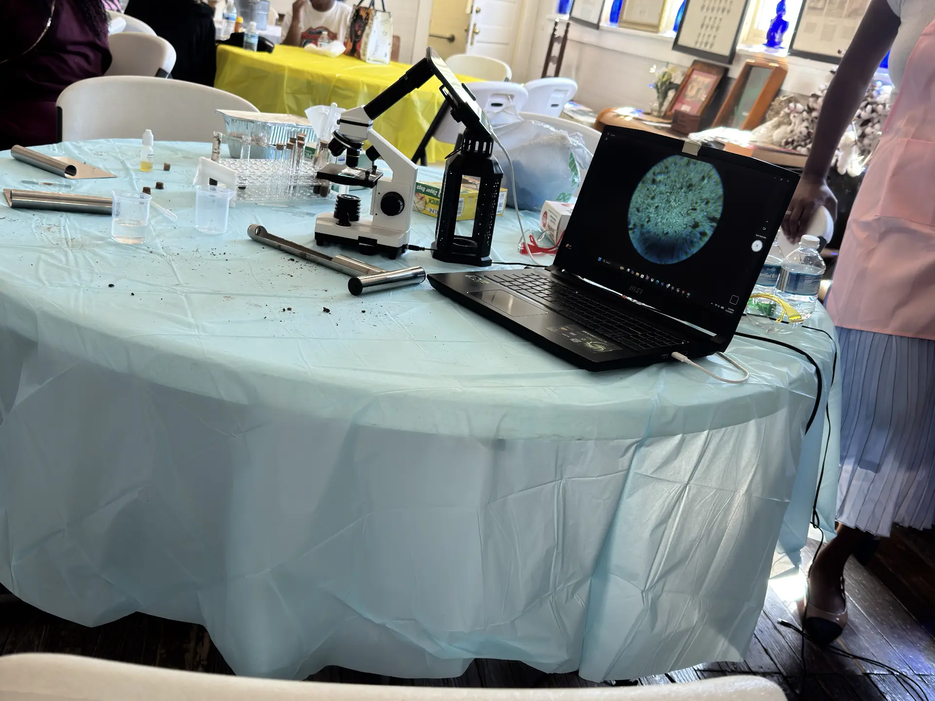 A microscope, test tubes and a laptop displaying a magnified specimen sit on a covered STEM Day work table.